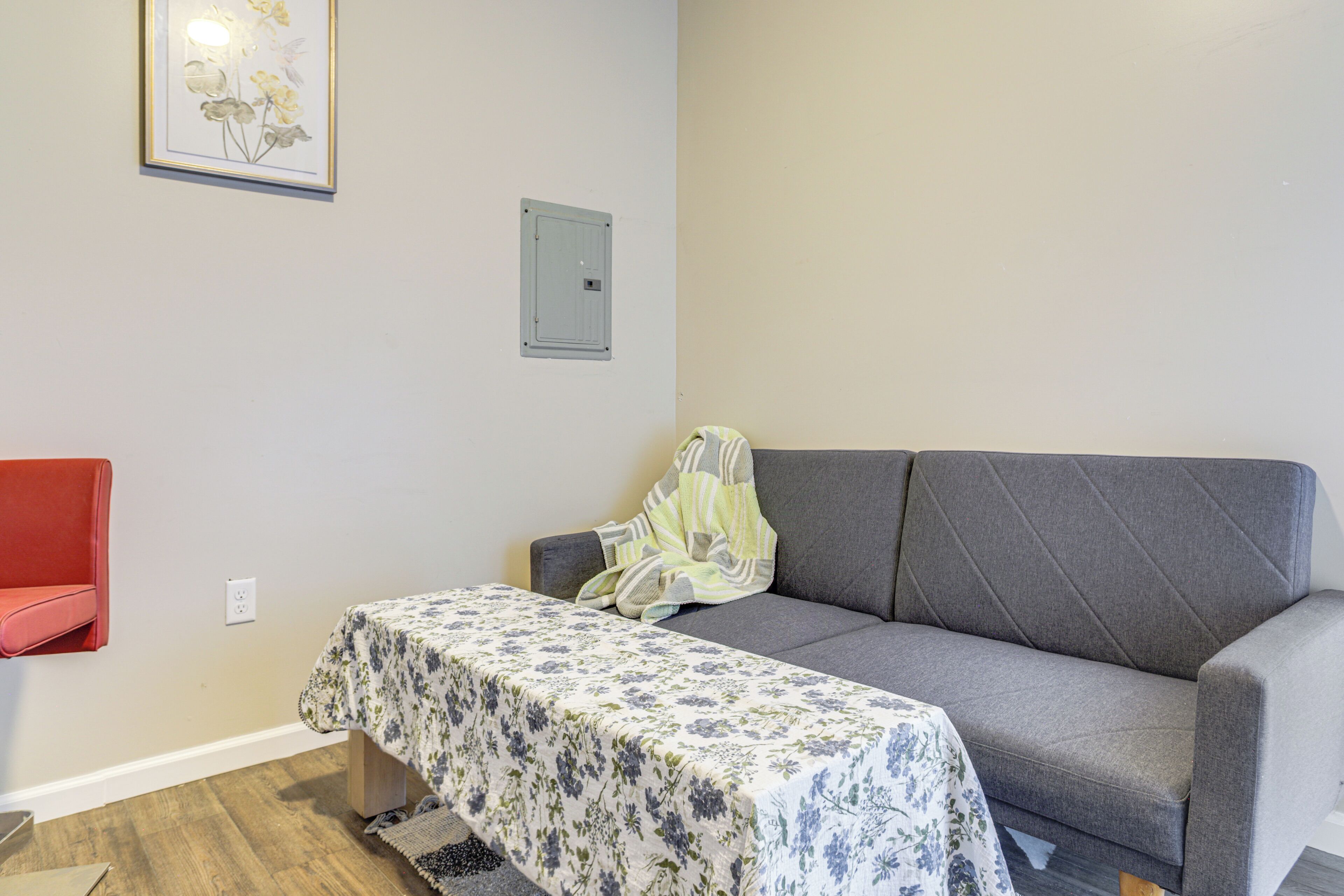Een kamer met een grijze bank, een klein tafeltje met een tafelkleed met bloemen, een rode stoel, en een ingelijste foto aan de muur.