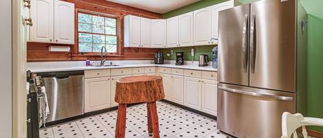 A kitchen with white cabinets, a stainless steel refrigerator, a wooden stool, and a tiled floor.