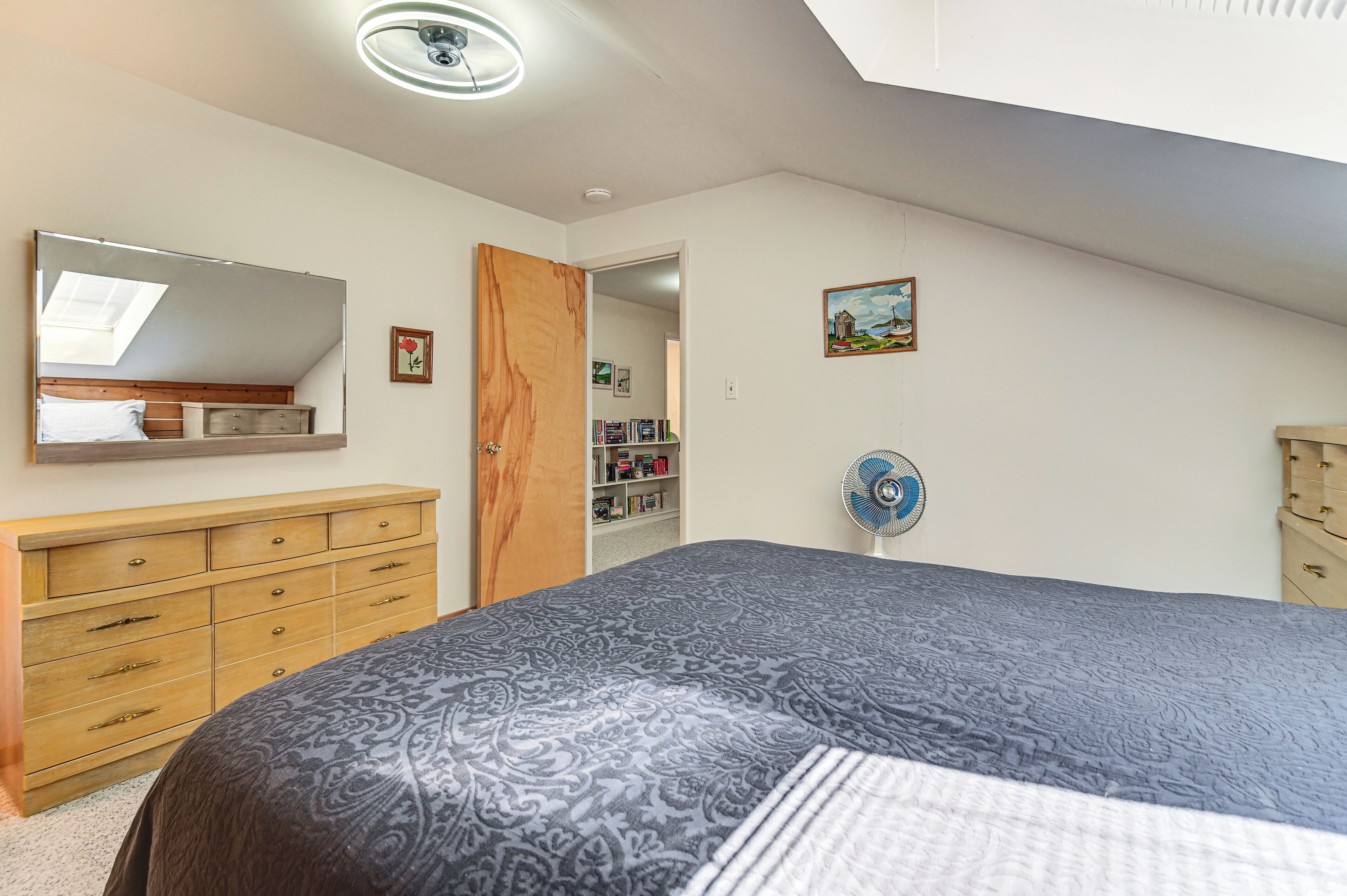 A bedroom with a large bed, a wooden dresser, a mirror, a fan, and a door leading to a closet.