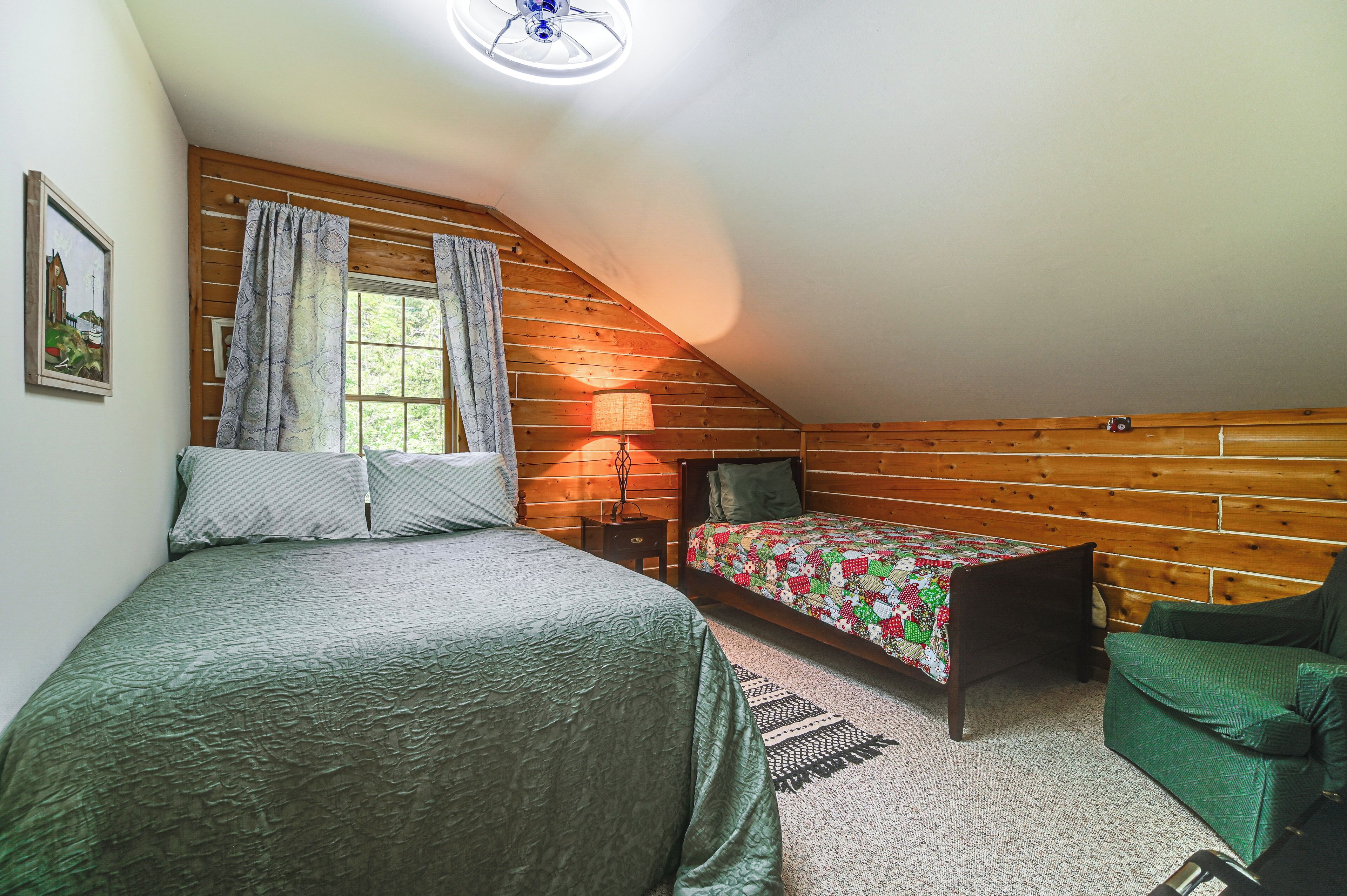 A cozy attic bedroom with a sloped ceiling, wooden walls, a large bed, a smaller bed, a chair, and a nightstand.