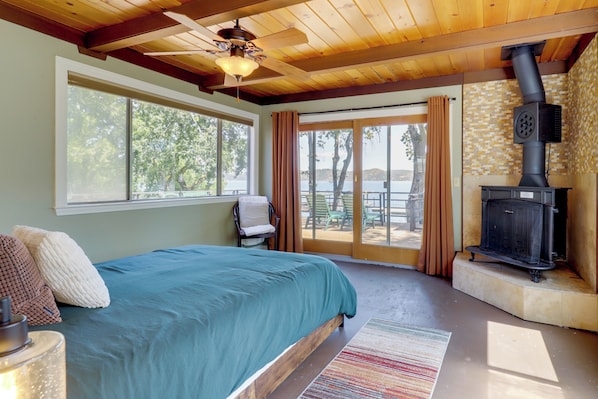 A bedroom with a large bed, a ceiling fan, a fireplace, and a view of the outdoors.