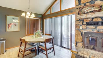 A dining area with a round table, chairs, a chandelier, a stone fireplace, and a window with blinds.