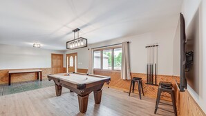 Een pooltafel in een ruimte met houten vloer, een bank, een televisie en een raam met gordijnen.