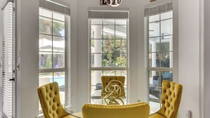 A room with a glass table and two yellow upholstered chairs, large windows with blinds, and a view of a pool and greenery outside.
