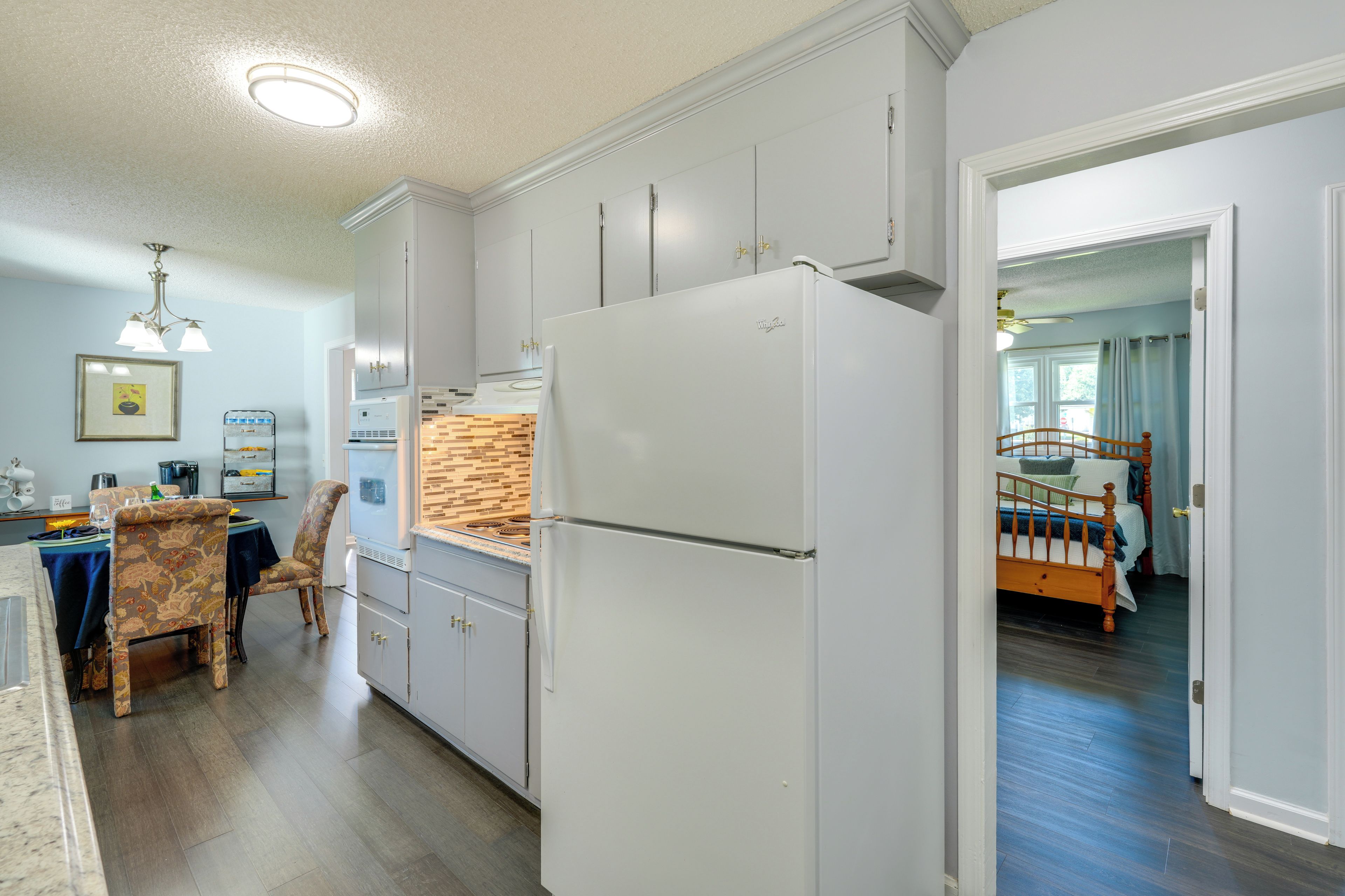 A modern kitchen with a built-in refrigerator, a dining area with a patterned table and chairs, and a view into a bedroom with a wooden bed frame.