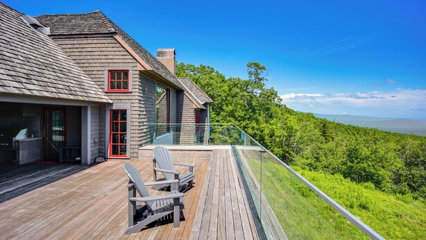 Een modern huis met een houten terras, een glazen balustrade en uitzicht op een weelderig groen landschap.