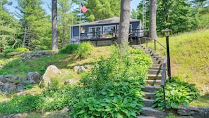 Une cabane dotée d’une terrasse et d’un escalier menant à celle-ci, entourée de verdure et d’arbres.