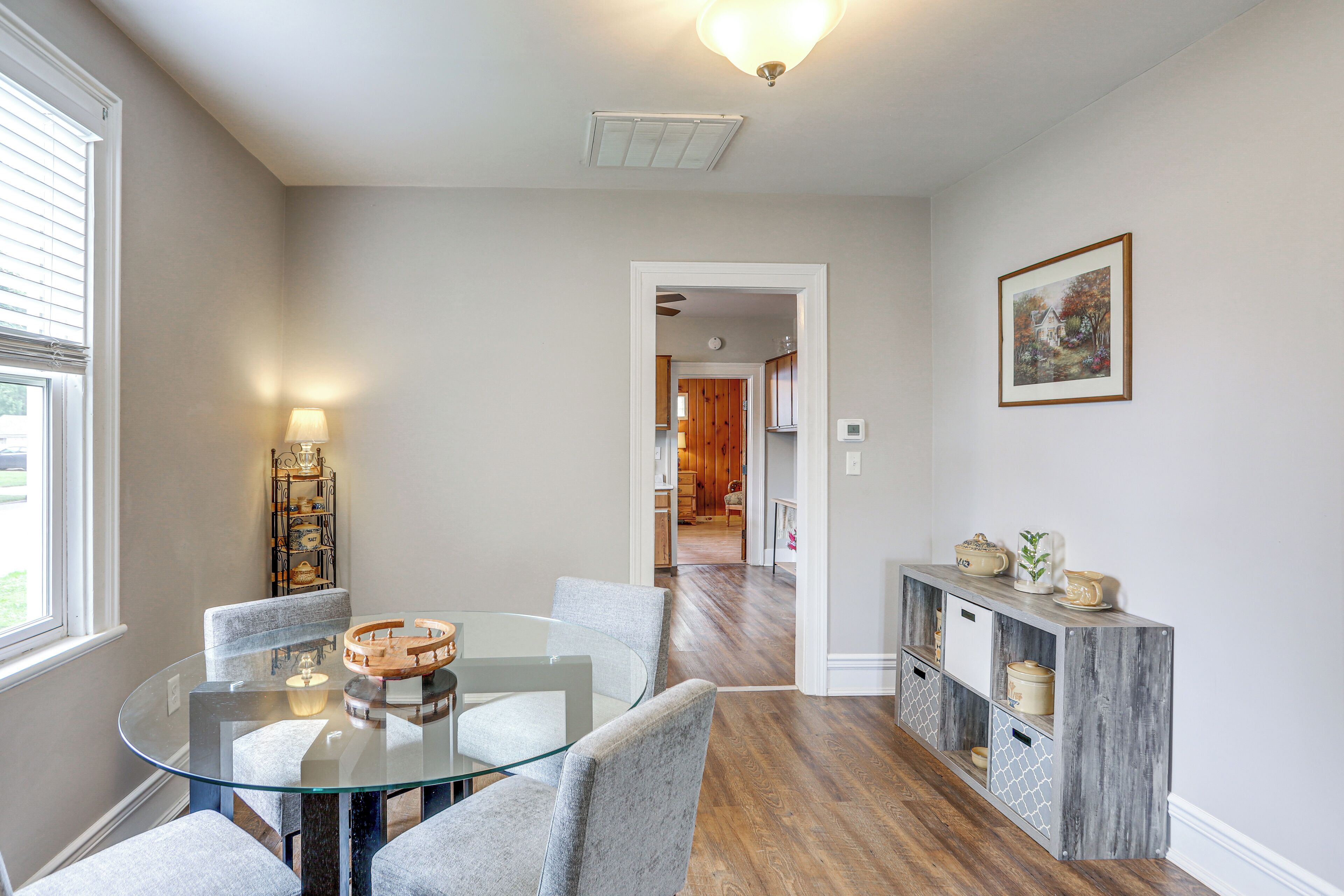 A dining area with a glass table and grey upholstered chairs, a wooden cabinet with storage, and a framed picture on the wall.