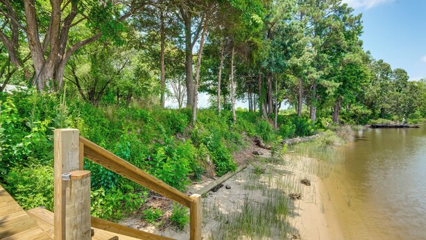 Eine Holzterrasse mit einer Treppe, die zu einer sandigen Fläche an einem ruhigen Gewässer führt, umgeben von dichtem Grün und Bäumen.
