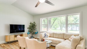 Ein modernes Wohnzimmer mit Fernseher, Sofa, Sesseln und einem großen Fenster.