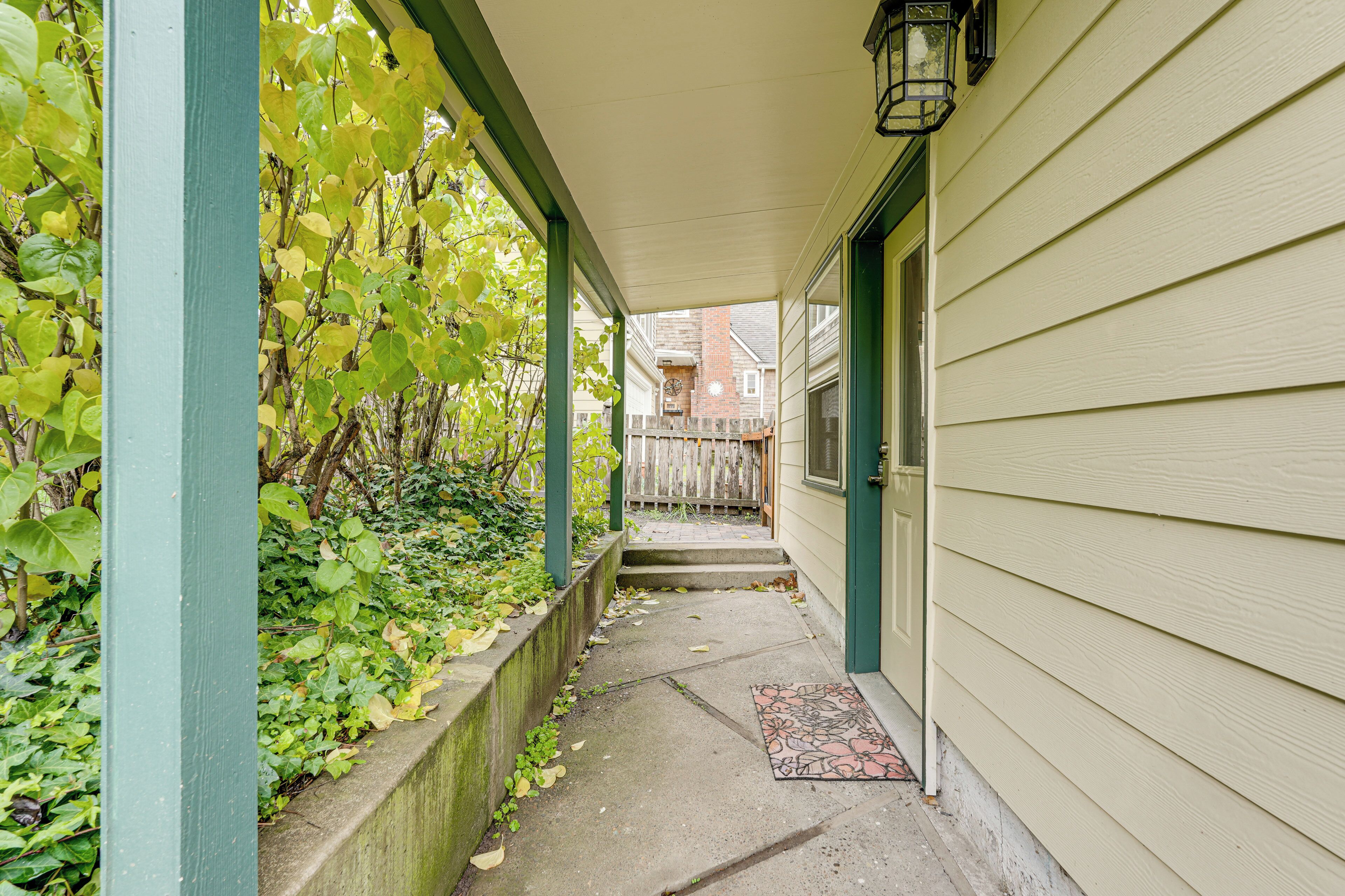 A covered porch with a concrete pathway, a wooden fence, and a brick building in the background.