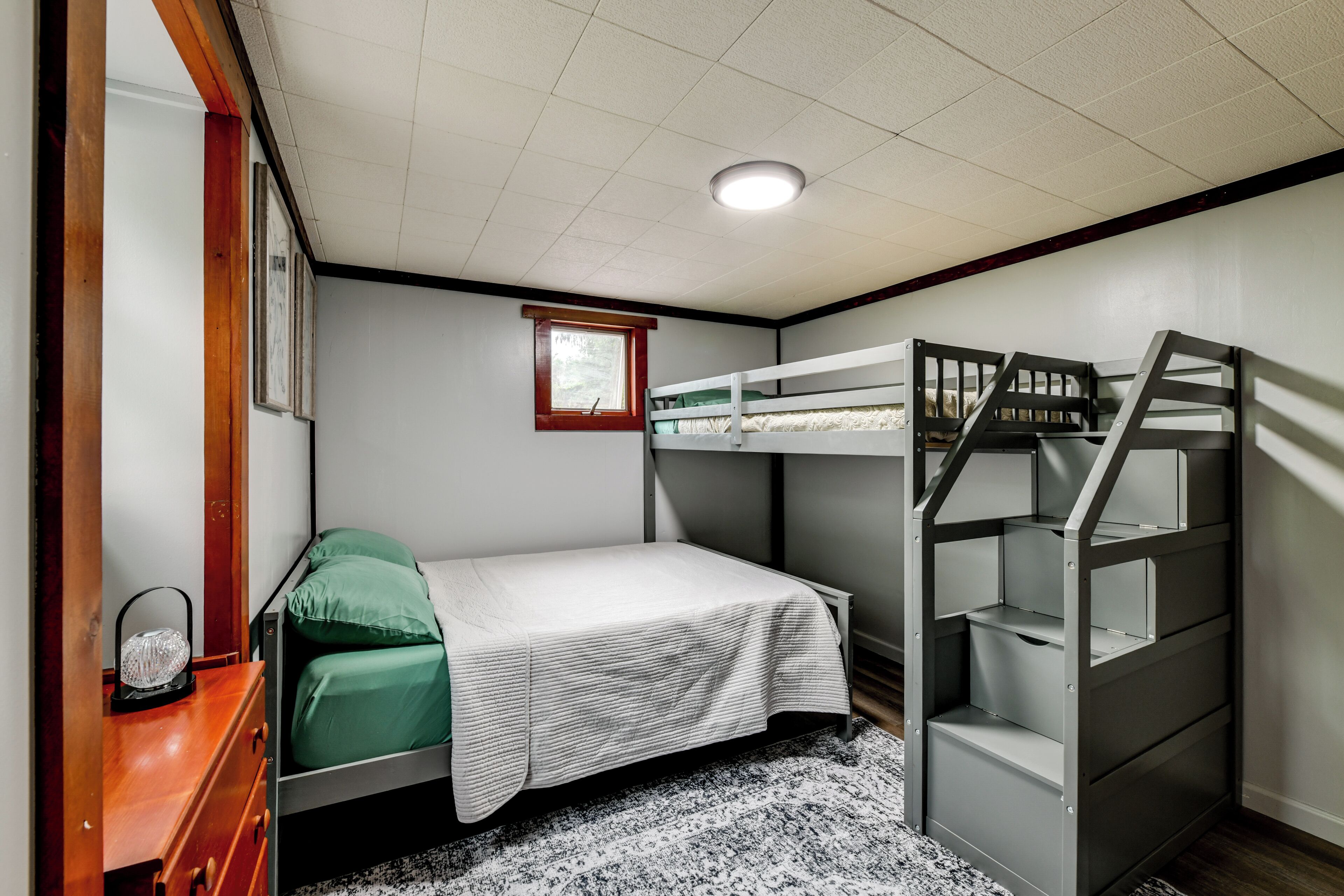 Une chambre avec des lits superposés, une commode en bois, une petite fenêtre et un luminaire au plafond.
