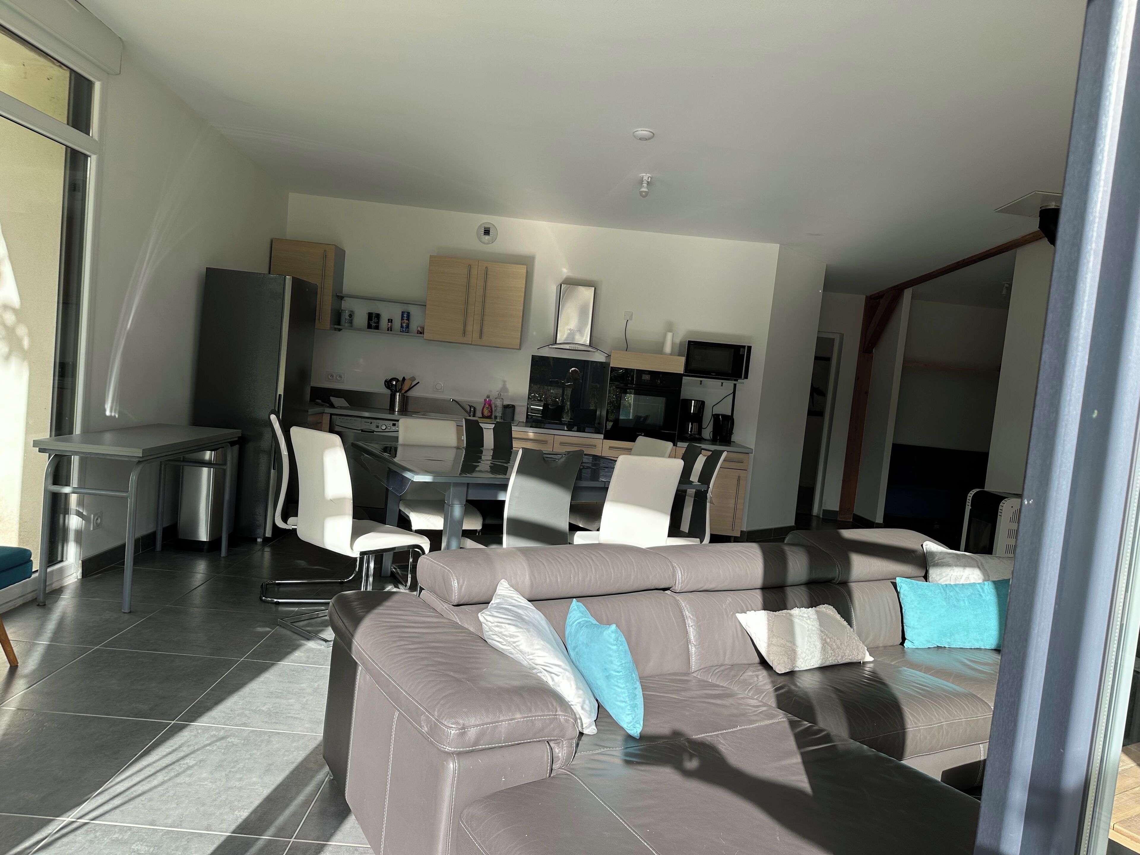 A modern living room with a grey leather sofa, a dining area with a glass table, and a kitchen with wooden cabinets and a built-in oven.