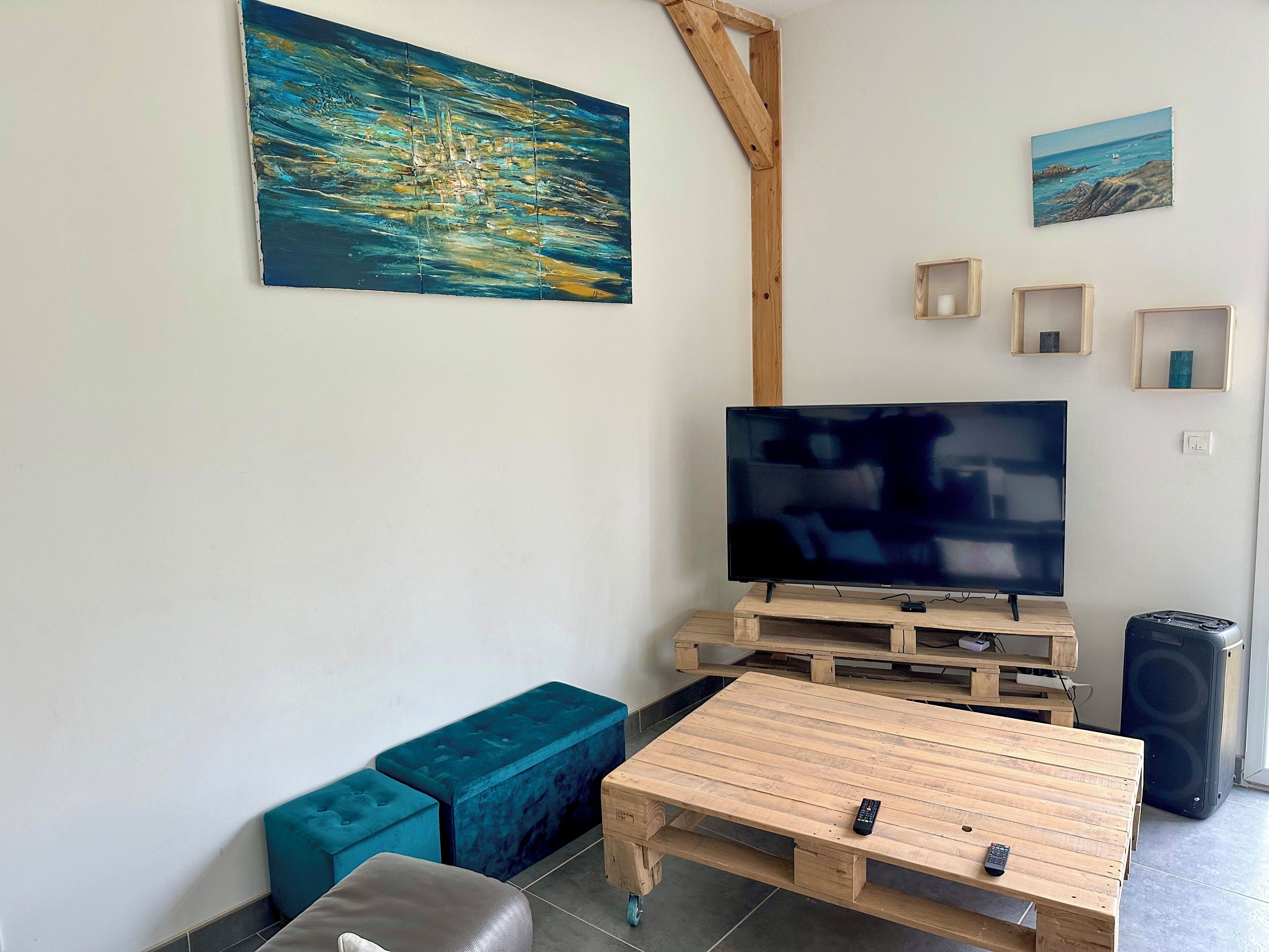 Una sala de estar con un televisor de pantalla plana grande, una mesa de centro de madera, un banco azul y un cuadro de una vista marina.