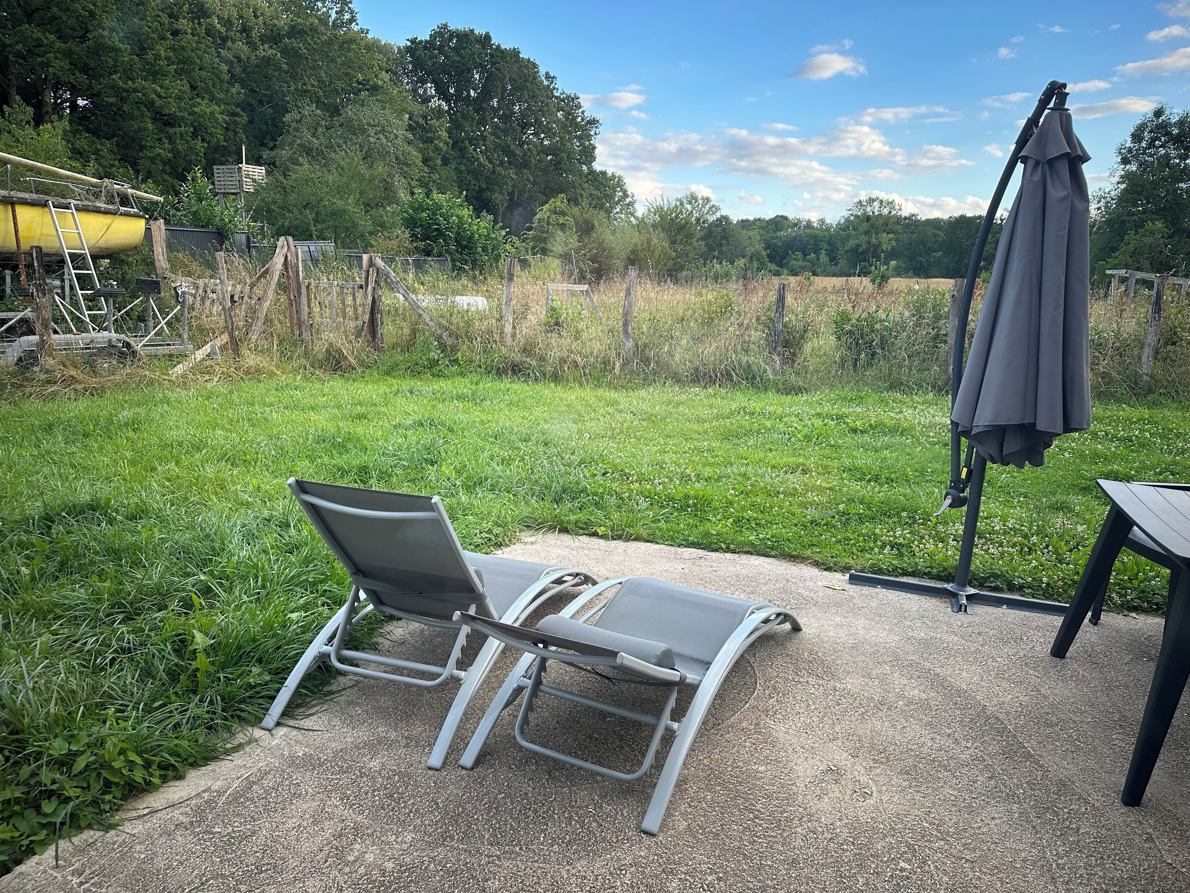 A patio area with a lounge chair and a table, surrounded by a grassy field and trees.