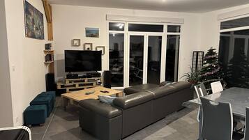 Een moderne woonkamer met een modulaire bank, een flatscreen televisie, een houten salontafel en een eethoek met een glazen tafel en stoelen.
