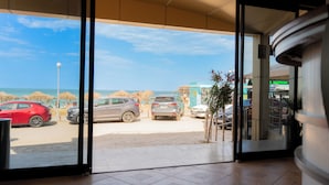 On aperçoit, à travers une porte vitrée, un parking avec plusieurs voitures, une plage et un bâtiment sur lequel on peut lire une enseigne.