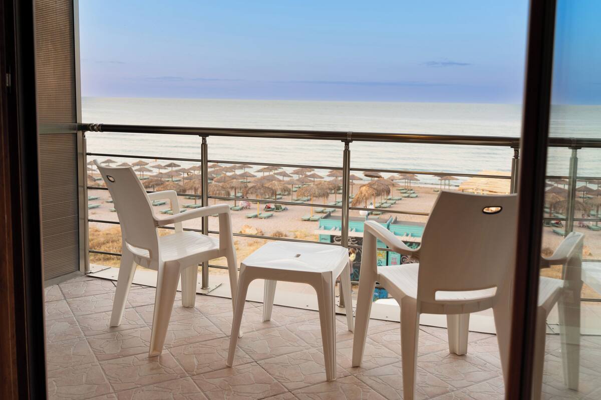 A balcony with two white plastic chairs and a small table, overlooking a beach with umbrellas and lounge chairs.