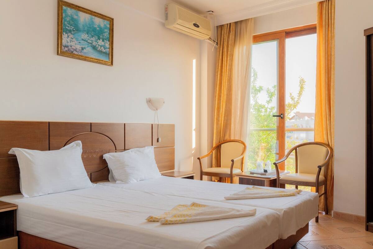 A hotel room with a bed, two chairs, a nightstand, a painting, and a window with curtains.