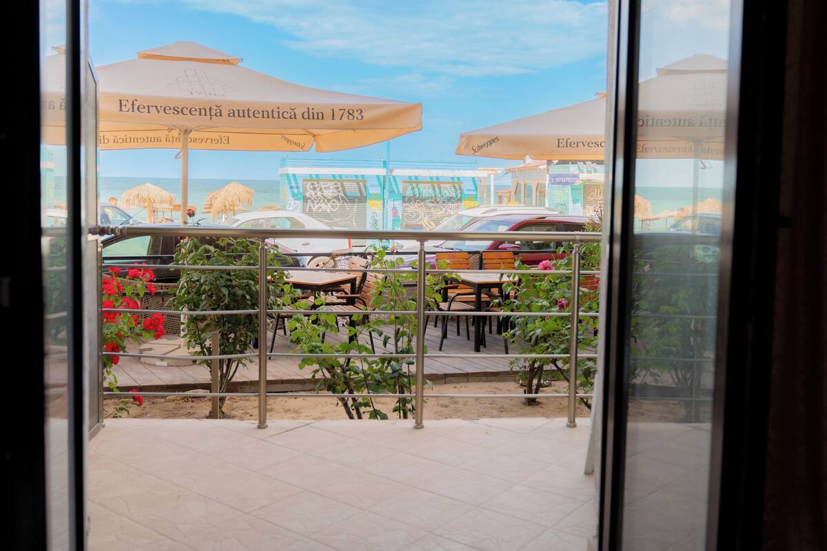 A view through a glass door of a restaurant patio with outdoor seating, umbrellas, and a view of the sea.