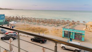 Une plage bordée de rangées de parasols et de chaises longues, quelques voitures garées à proximité, et un bâtiment couvert de graffitis.