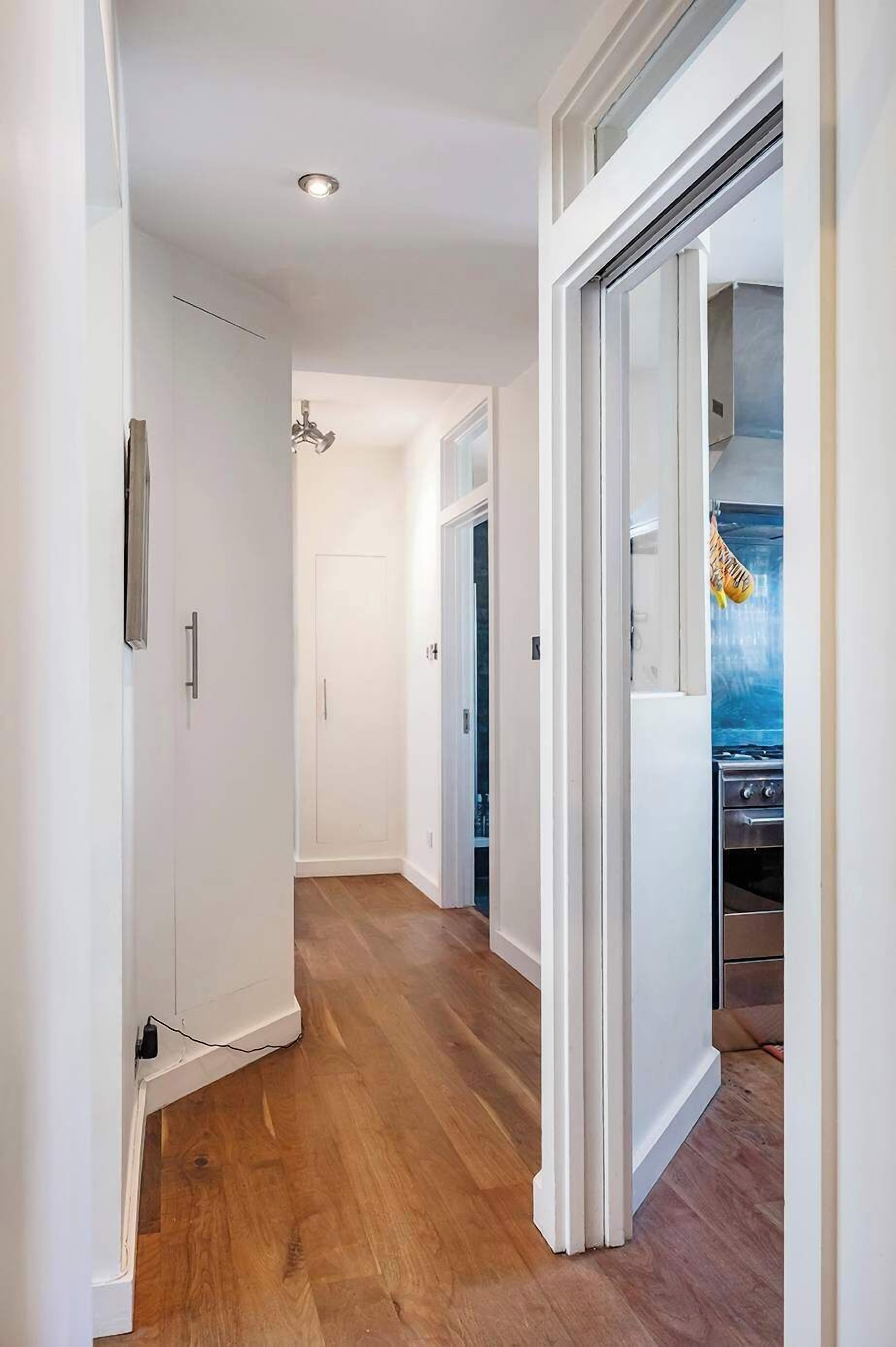 A narrow hallway with wooden flooring, white walls, and a kitchen area visible through an open door.
