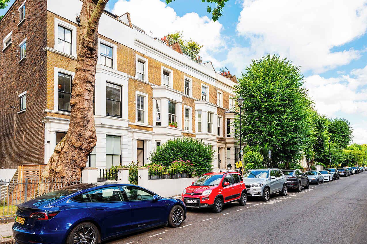 A row of parked cars along a tree-lined street with a row of terraced houses.