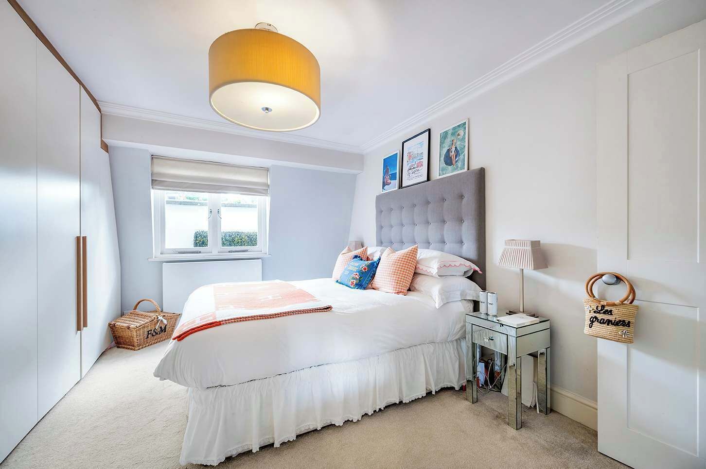 A bedroom with a large bed, a bedside table, a lamp, a basket, and a window.