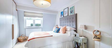 A bedroom with a large bed, a bedside table, a lamp, a basket, and a window.
