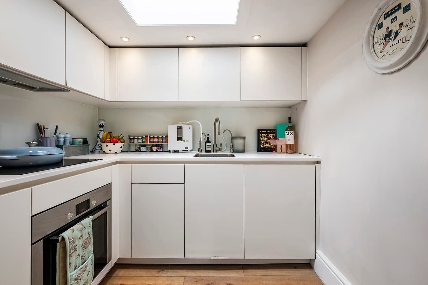 A modern kitchen with white cabinets, a built-in oven, a microwave, and a sink. There is a window above the sink and a clock on the wall.