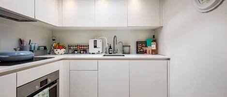 A modern kitchen with white cabinets, a built-in oven, a microwave, and a sink. There is a window above the sink and a clock on the wall.