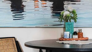 A room with a painting of two people in water, a small table with a vase and bottles, and a chair with a striped cushion. - The Light Chambers Notting Hill- by Out of Office Lifestyle (London)