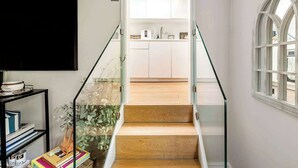 A staircase with glass railings leading to a kitchen area with white cabinets and a window with a white frame. - The Light Chambers Notting Hill- by Out of Office Lifestyle (London)
