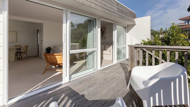 Een modern huis met een houten terras en schuiframen die uitkomen op een interieur met een eethoek, een bank en een stoel.