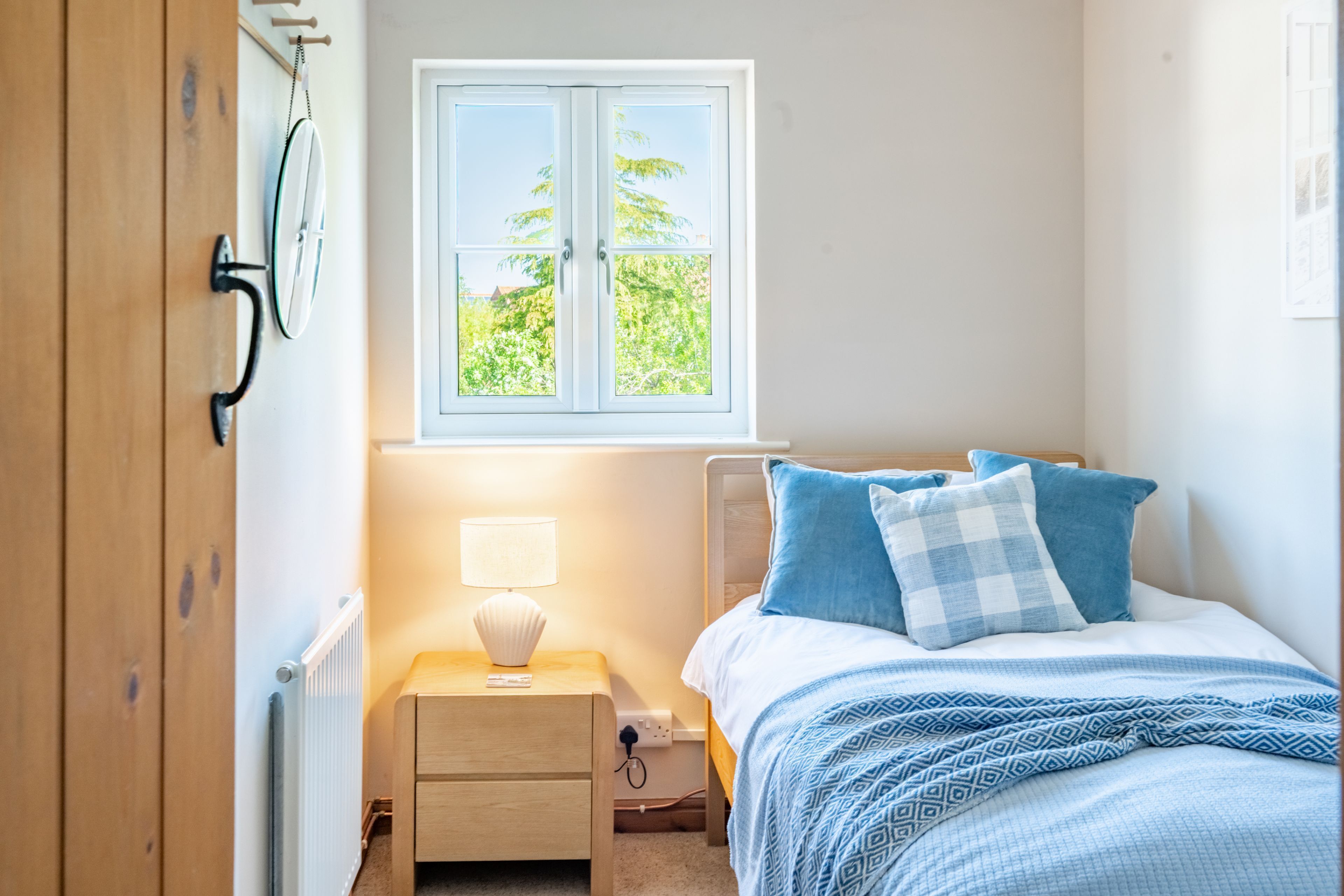 A bedroom with a bed, a nightstand, a lamp, a window with a view of greenery, and a wooden wardrobe.