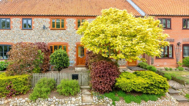 Ein Haus aus Stein und Backstein mit einem Ziegeldach, einem Holzzaun und verschiedenen Sträuchern und Bäumen im Garten.