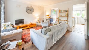 Ein Wohnzimmer mit einer Couch, einem Sessel und einem Fernseher. Außerdem ist ein Bücherregal und eine Lampe zu sehen.