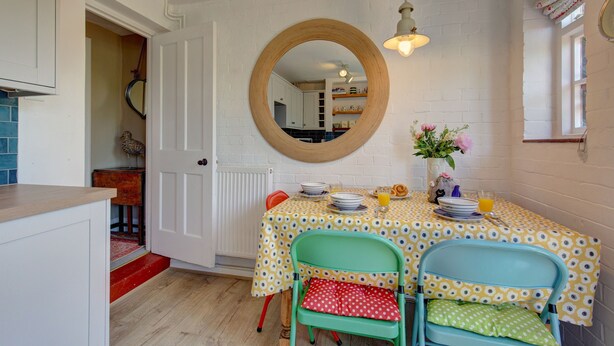 Een zithoek met een ronde spiegel, een tafel met een tafelkleed met gele stippen, en twee stoelen met rode en groene kussens.