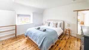 Une chambre avec un plancher en bois, un lit recouvert d’une couverture bleue, un radiateur, une fenêtre donnant sur un paysage agréable et un tabouret en bois surmonté d’une lampe.