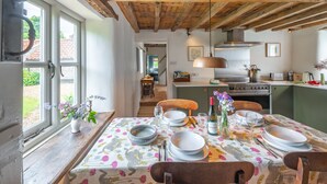Un coin repas avec une table dressée pour quatre personnes, des poutres en bois au plafond et, au fond, une vue sur la cuisine.