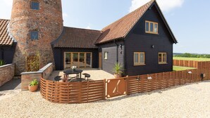Une grande maison avec une tour en briques, une façade sombre et une terrasse en gravier aménagée avec du mobilier d’extérieur.