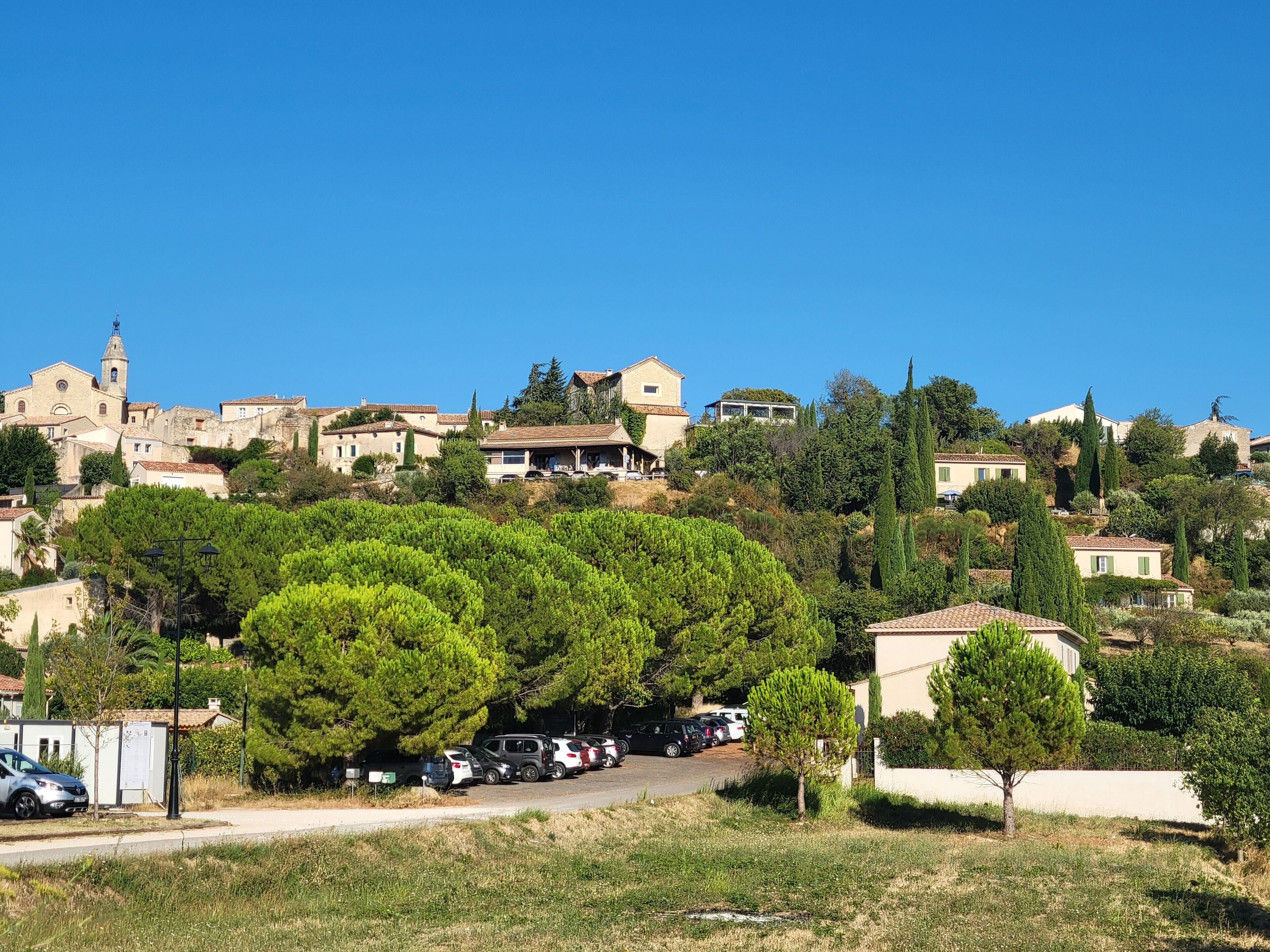A hillside village with traditional stone buildings, a church with a bell tower, and parked cars along a road.