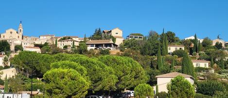 A hillside village with traditional stone buildings, a church with a bell tower, and parked cars along a road.