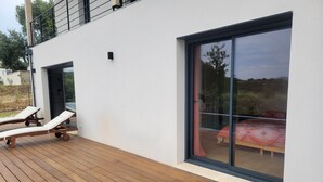 Um deck moderno ao ar livre com duas poltronas de descanso e uma porta de vidro deslizante que dá para um quarto com colcha vermelha estampada.