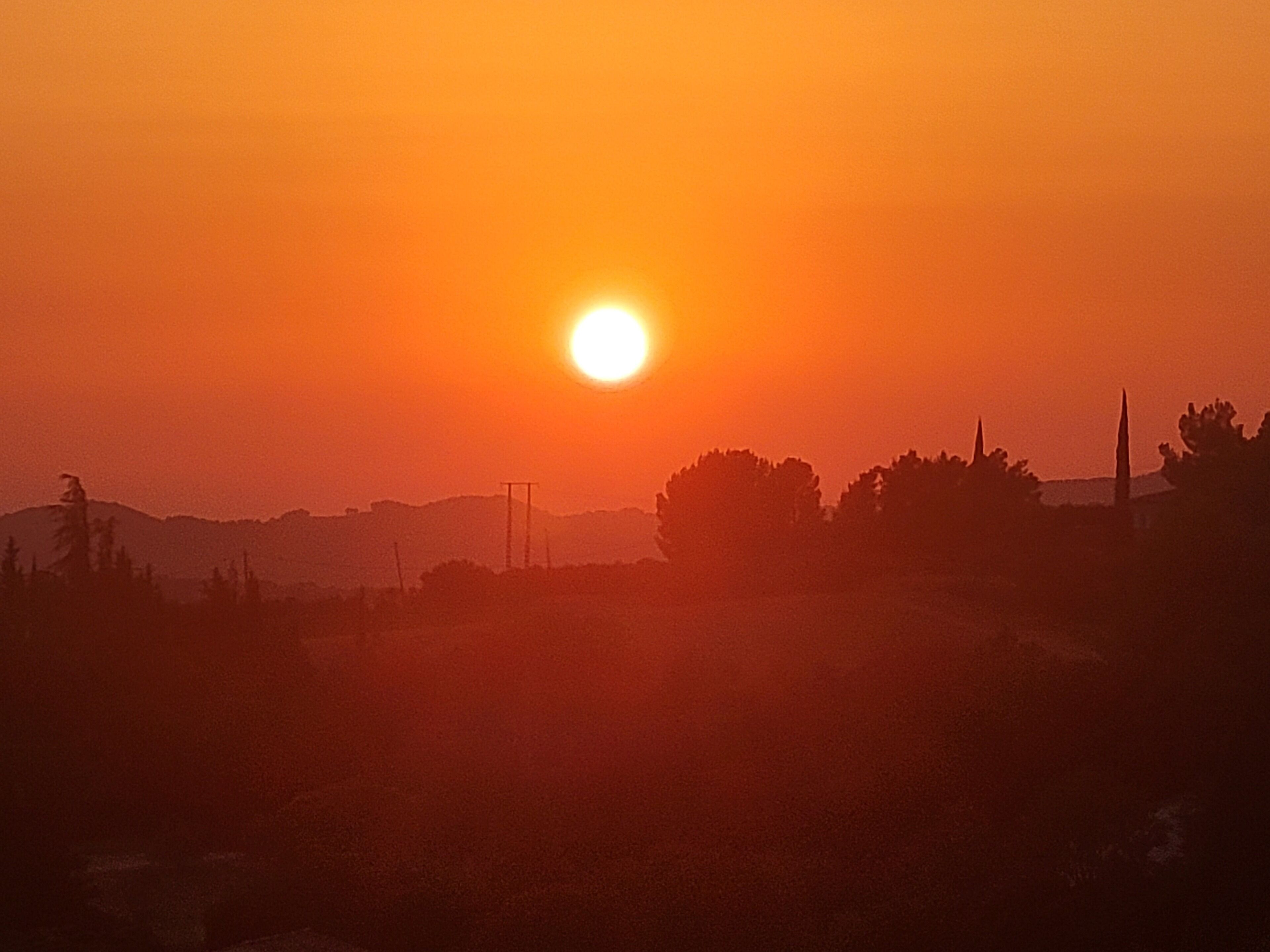 Ein Sonnenuntergang mit tiefstehender Sonne, die Bäume und ferne Hügel silhouettiert.