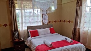Un lit bien fait, avec une literie blanche et rouge, une table de chevet avec une lampe et une fenêtre ornée de rideaux légers.