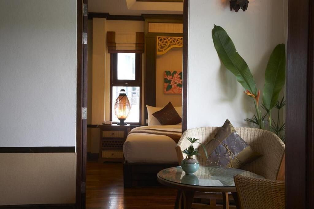 Una habitación con una cama, una mesita con un florero y una planta grande.