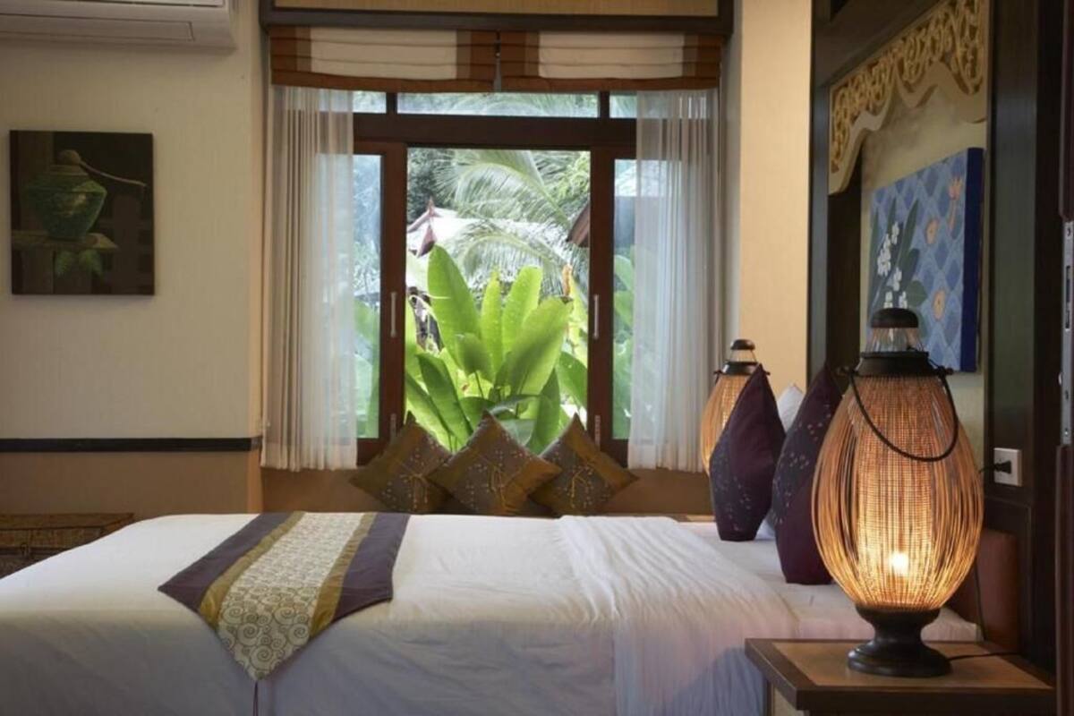 Quarto de hotel com cama, uma janela grande com cortinas, luminária decorativa e um quadro na parede.