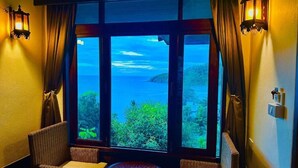 Ein Zimmer mit großem Fenster und Blick auf ein Gewässer, zwei Korbstühle, ein kleiner runder Tisch und Wandleuchten.