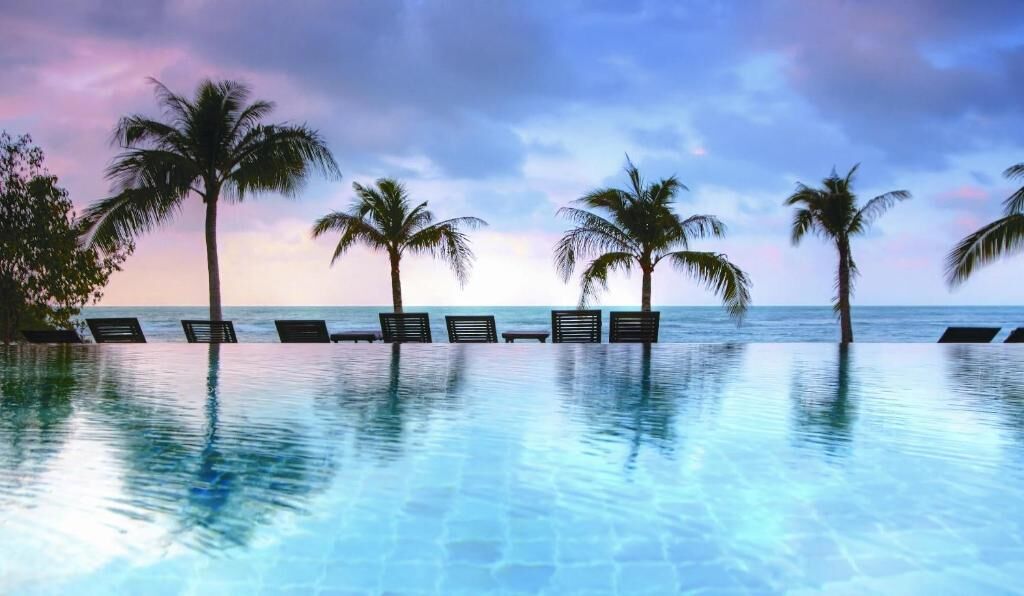 Área de piscina com espreguiçadeiras e palmeiras, com vista para o oceano.